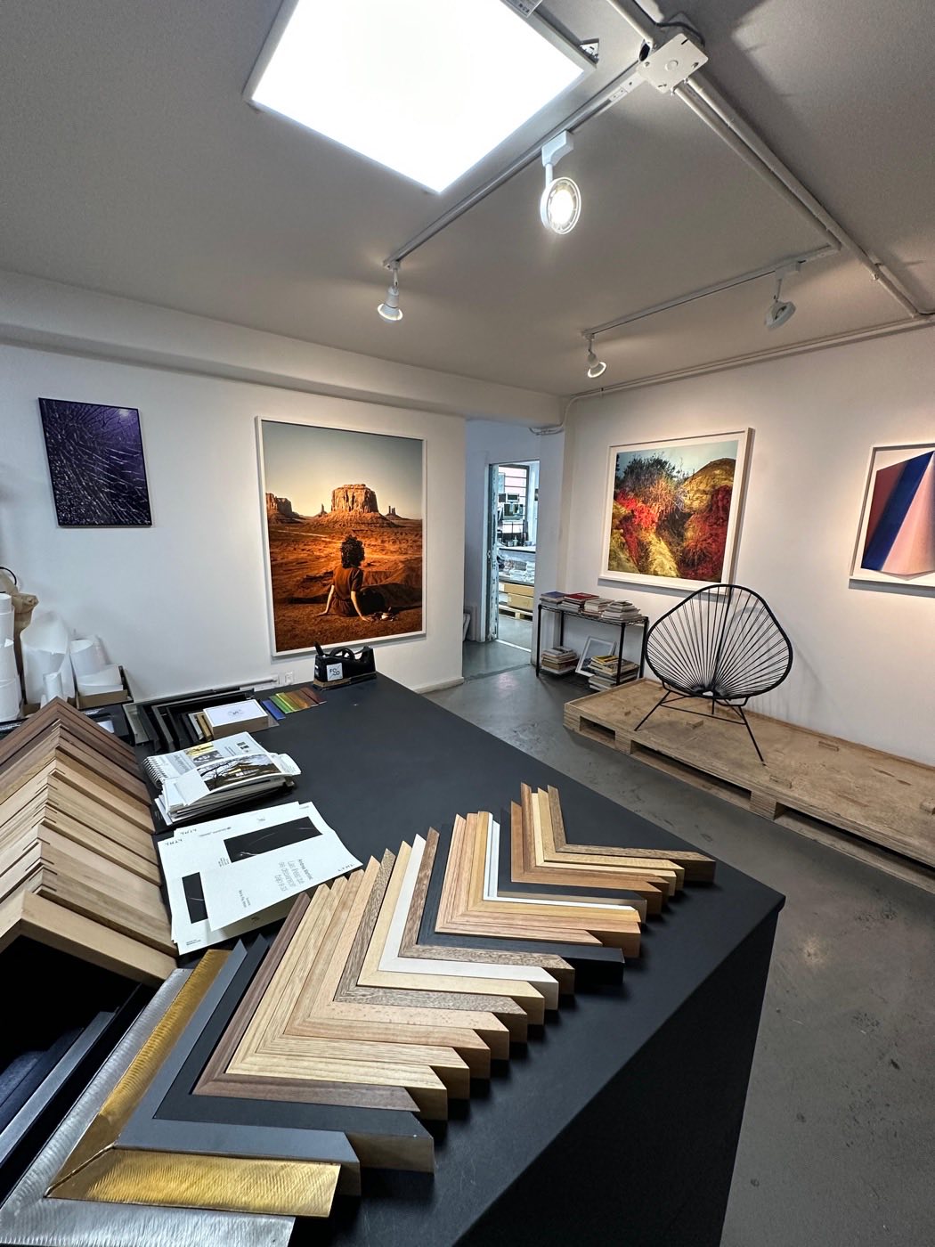 Atelier showroom with hung work and a table covered in frame samples — natural wood, ebony, walnut, and metallic finishes